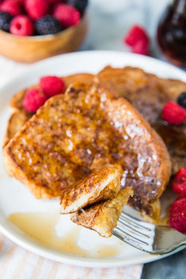 several slices of vegan french toast on a white plate topped with syrup, raspberries and blackberries. the focus is on a fork where a bite of the french toast is on