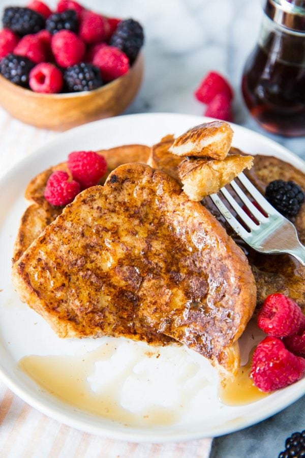 several slices of vegan french toast on a white plate topped with syrup, raspberries and blackberries. a bite has been taken out of on the the pieces