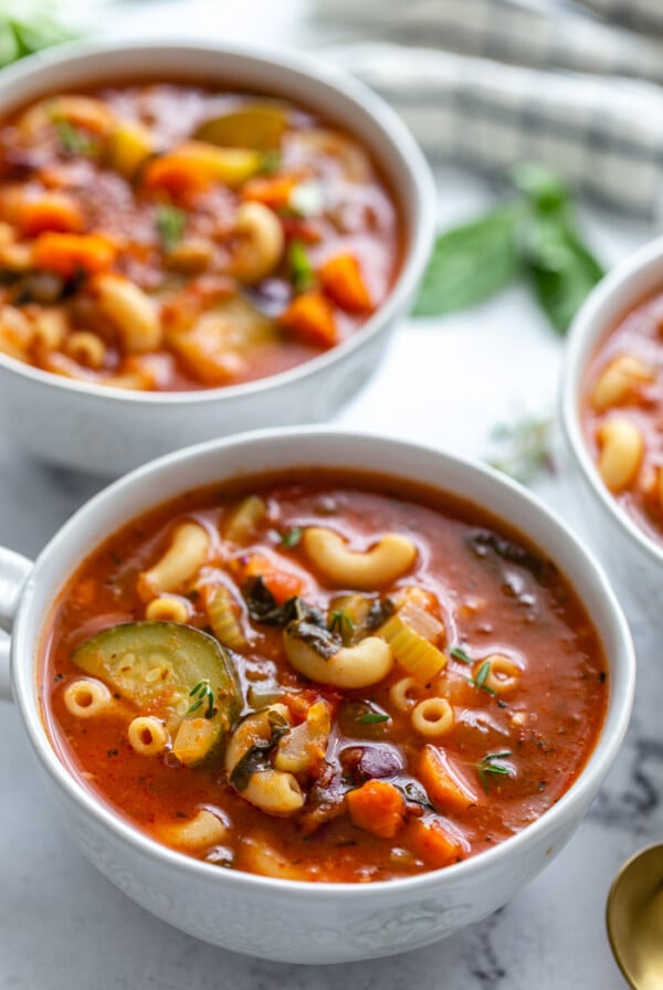 several bowls of vegetable minestrone soup in soup mugs with fresh herbs in the background