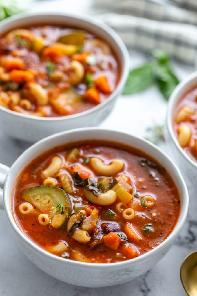 several bowls of vegetable minestrone soup in soup mugs with fresh herbs in the background