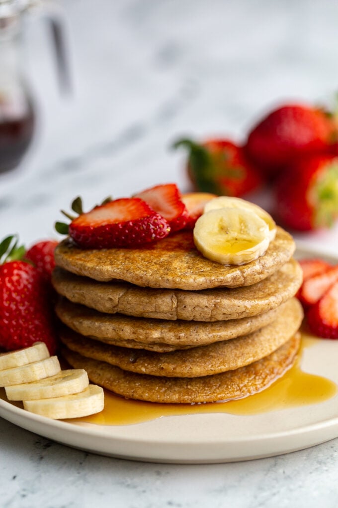 stack of oat flour pancakes on a plate that are topped with banana and strawberry slices and maple syrup. More banana and strawberries on the side of the plate