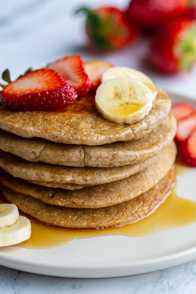 stack of oat flour pancakes on a plate that are topped with banana and strawberry slices and maple syrup. More banana and strawberries on the side of the plate