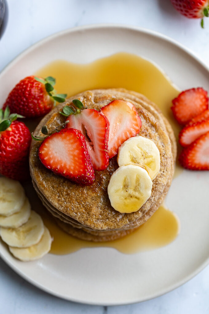 stack of oat flour pancakes on a plate that are topped with banana and strawberry slices and maple syrup. More banana and strawberries on the side of the plate