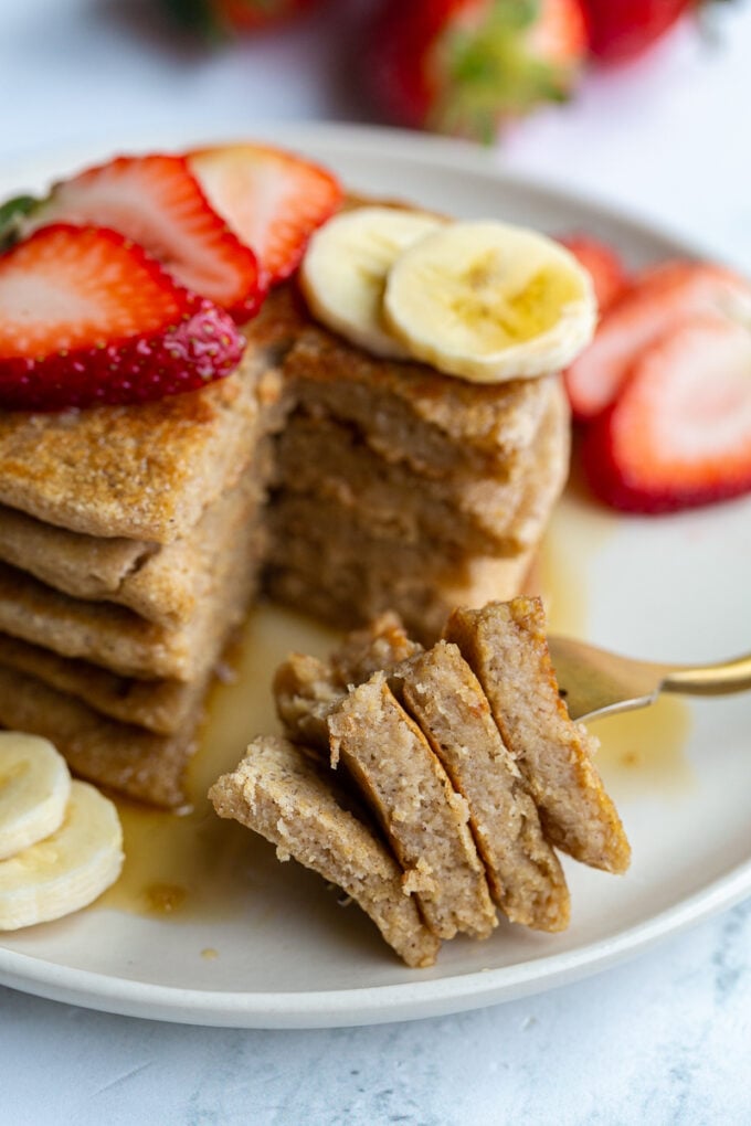 stack of oat flour pancakes on a plate that are topped with banana and strawberry slices and maple syrup. More banana and strawberries on the side of the plate. A bite has been taken out of the pancakes and the stacked bite of pancakes is resting on the plate