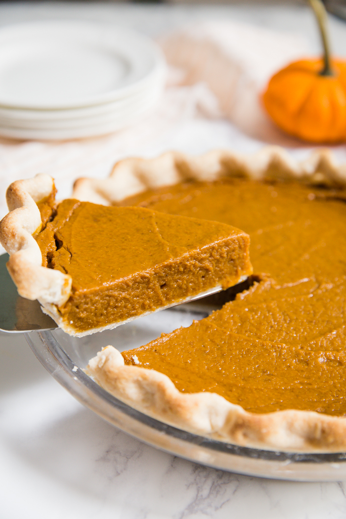 slice of vegan pumpkin pie being lifted with the rest of the pie. orange pumpkin in the background