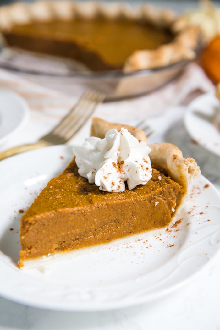 slice of vegan pumpkin pie on a white plate with whipped cream and cinnamon on top. Full pie in the background