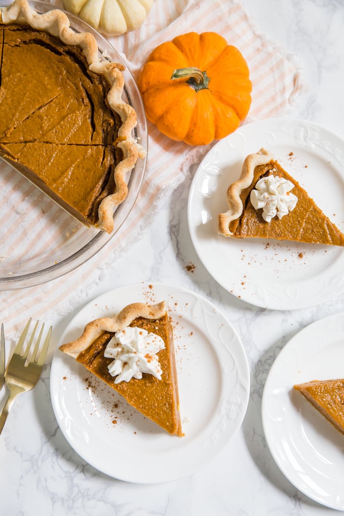 top down shot of vegan pumpkin pie slices on white plates and the full pie in the corner with a fresh pumpkin next to it