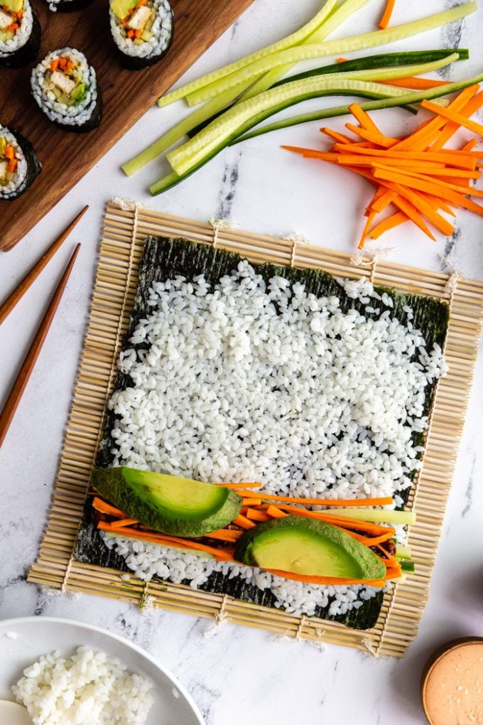 sushi being made on a marble board. A sushi mat has an unrolled roll of sushi on it that's topped with rice, cucumber, carrot, and avocado