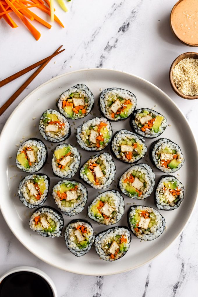 top down shot of a plate of tofu sushi filled with tofu, carrots, avocado, and cucumber. fresh veggies in one corner, soy sauce in another corner, sesame seeds and spicy mayo in another corner. all on a marble board