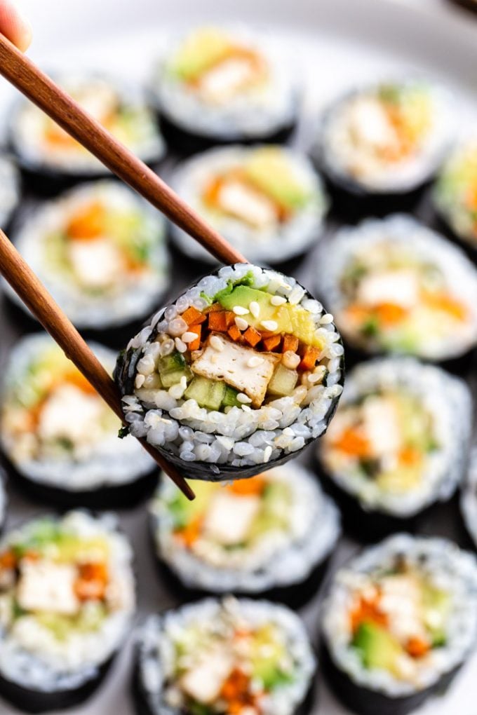 shot of a plate of tofu sushi filled with tofu, carrots, avocado, and cucumber. one piece of sushi is being held up by chopsticks