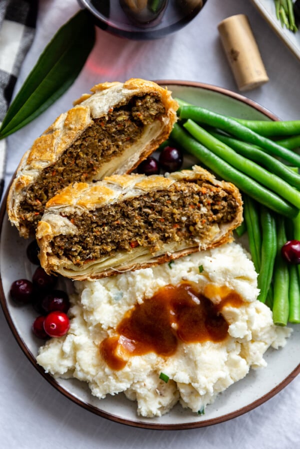 plate filled with 2 slices of vegan wellington, mashed potatoes and gravy and well as green beans. glass of wine in the corner of the shot