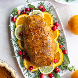 vegan turkey loaf sitting on a bed of fresh herbs and citrus slices. plates of green beans, pumpkin pie, and mashed potatoes are in the corner of the shot