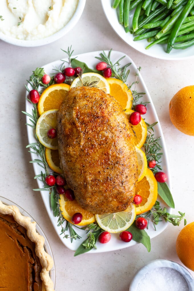 vegan turkey loaf sitting on a bed of fresh herbs and citrus slices. plates of green beans, pumpkin pie, and mashed potatoes are in the corner of the shot