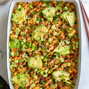 vegetarian sushi bake inside of a casserole dish that's topped with heaps of avocado, sesame seeds and drizzled with red sriracha, orange spicy mayo, and kewpie mayo. It's also sprinkled with chopped green onion