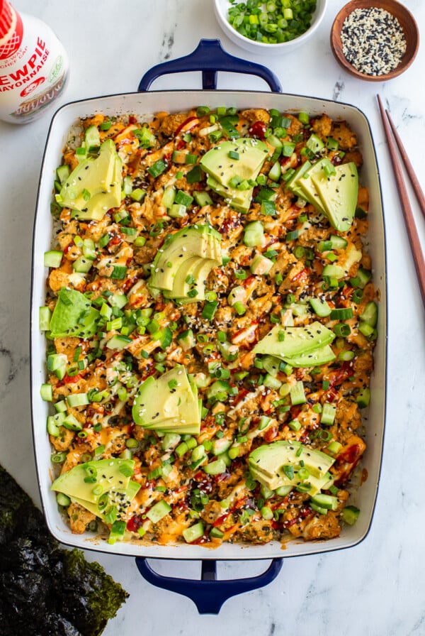 vegetarian sushi bake inside of a casserole dish that's topped with heaps of avocado, sesame seeds and drizzled with red sriracha, orange spicy mayo, and kewpie mayo. It's also sprinkled with chopped green onion