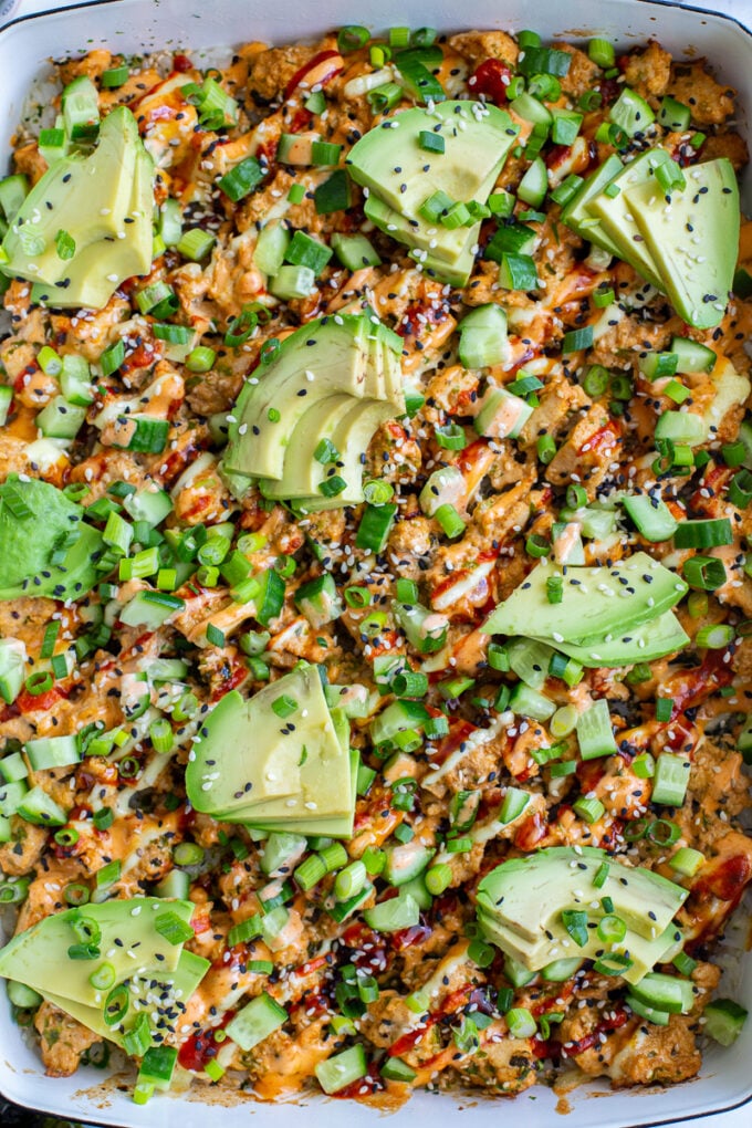 vegetarian sushi bake inside of a casserole dish that's topped with heaps of avocado, sesame seeds and drizzled with red sriracha, orange spicy mayo, and kewpie mayo. It's also sprinkled with chopped green onion