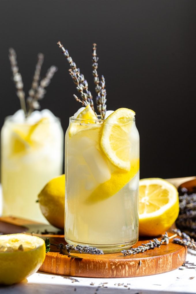 glass of vodka lemonade with slices of lemon and lavender in it. sitting on a wood board with a black background