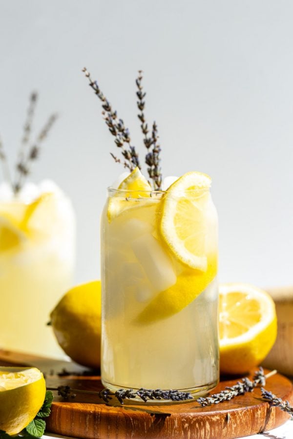 glass of lemonade with slices of lemon and lavender in it. sitting on a wood board with a white background