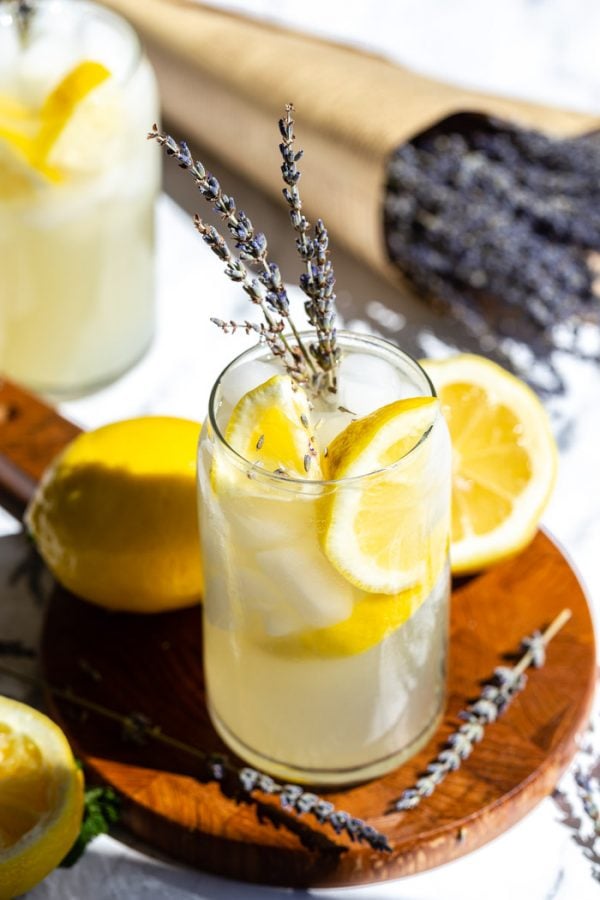 glass of lemonade with slices of lemon and lavender in it. sitting on a wood board with lemons and lavender all around it