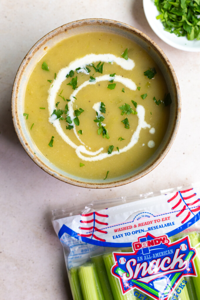 celery soup in a bowl topped with a little drizzle of cream. there's a bag od dandy celery in the corner of the shot