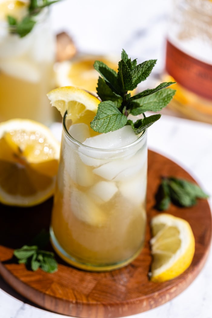 yellow/ orange whiskey cocktail on a wood board with lemon and mint popping out the top and a bottle of bourbon in the background