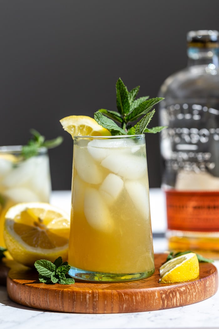 yellow/ orange whiskey cocktail on a wood board with lemon and mint popping out the top and a bottle of bourbon in the background