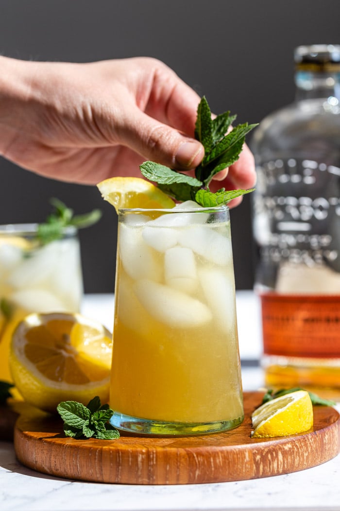 yellow/ orange whiskey cocktail on a wood board with lemon popping out the top and a bottle of bourbon in the background. a hand place a sprig of mint into the cocktail