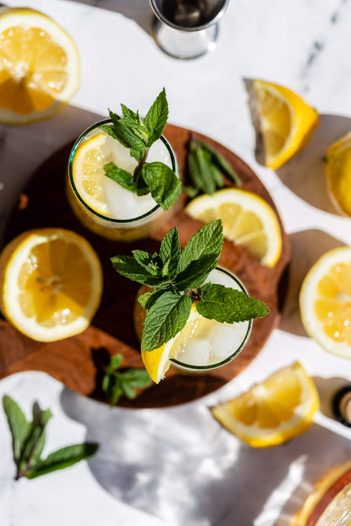 top down shot of 2 whiskey cocktails with lemon and mint all around on a round wood board